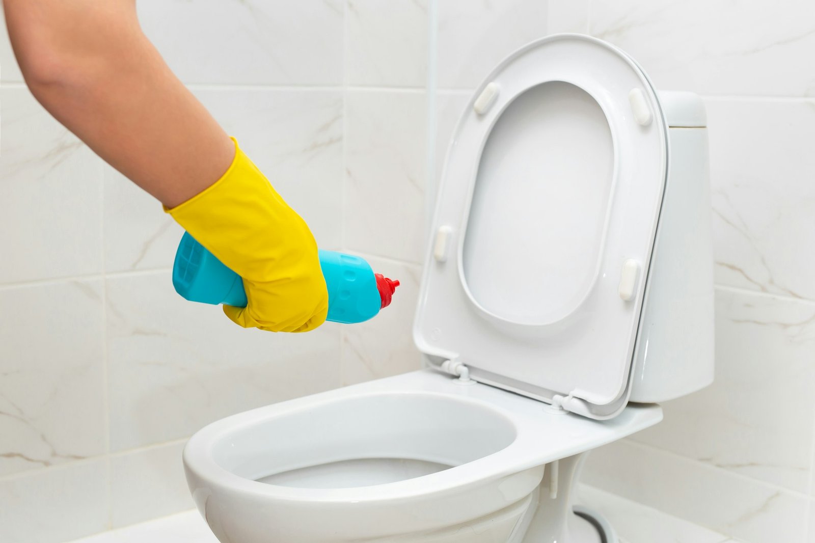 Cleaner in yellow rubber gloves pouring detergent under rim of toilet closeup