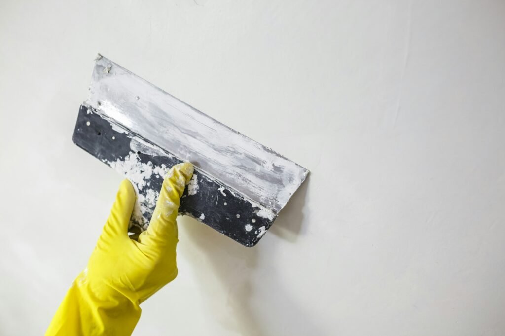 worker's hand in yellow gloves holding putty knife patching a hole with spatula